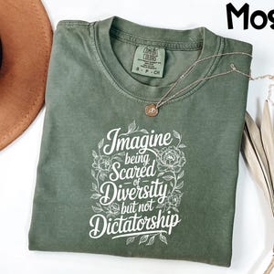May include: A moss green t-shirt with the text "Imagine being scared of Diversity but not Dictatorship" in white script. The shirt has a crew neck and short sleeves. A gold necklace with a pendant is draped across the shirt.