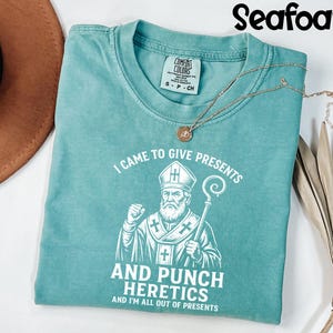 May include: Seafoam green t-shirt with the text "I CAME TO GIVE PRESENTS AND PUNCH HERETICS AND I'M ALL OUT OF PRESENTS" and a graphic of a bishop. A gold necklace with a pendant is on the shirt.