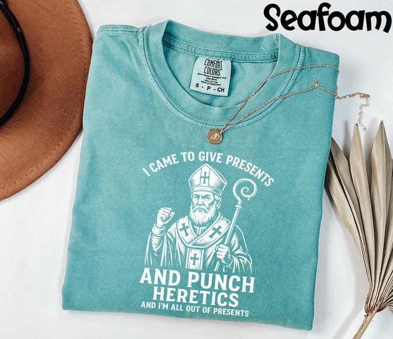 May include: Seafoam green t-shirt with the text "I CAME TO GIVE PRESENTS AND PUNCH HERETICS AND I'M ALL OUT OF PRESENTS" and a graphic of a bishop. A gold necklace with a pendant is on the shirt.