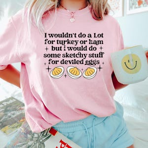 May include: A light pink t-shirt with the text "I wouldn't do a Lot for turkey or ham + but I would do + some sketchy stuff for deviled eggs +" and a graphic of deviled eggs. A white mug with a yellow smiley face is held in the hand.