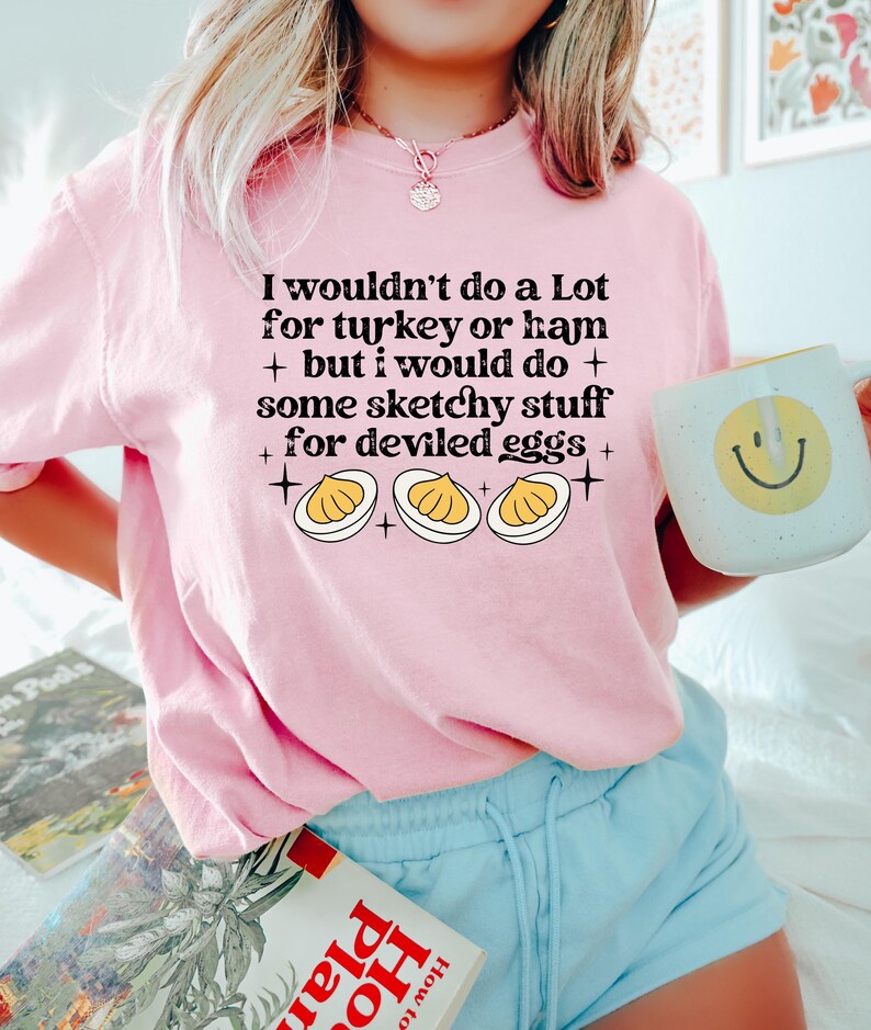 May include: A light pink t-shirt with the text "I wouldn't do a Lot for turkey or ham + but I would do + some sketchy stuff for deviled eggs +" and a graphic of deviled eggs. A white mug with a yellow smiley face is held in the hand.