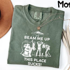 May include: Moss green t-shirt with the text "BEAM ME UP THIS PLACE SUCKS!" and a graphic of a retro woman, people, and a UFO. The shirt has a crew neck and short sleeves. A gold necklace with a pendant is draped across the shirt.