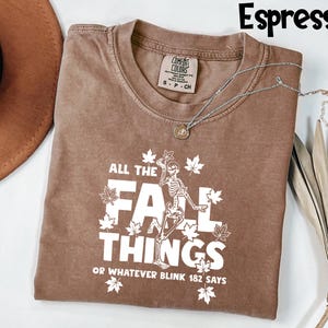 May include: Espresso-colored t-shirt with the text "ALL THE FALL THINGS OR WHATEVER BLINK 182 SAYS" in white. The shirt features a skeleton graphic and maple leaf designs. A silver necklace with a pendant is draped across the shirt.