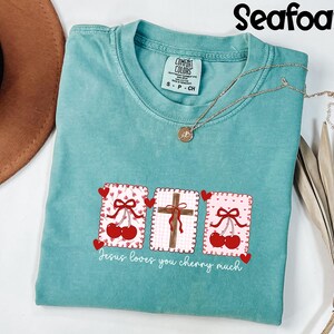May include: Seafoam green t-shirt with a graphic design featuring cherries, a cross, and the text "Jesus loves you cherry much." The shirt has a crew neck and short sleeves. A gold necklace is draped across the shirt.