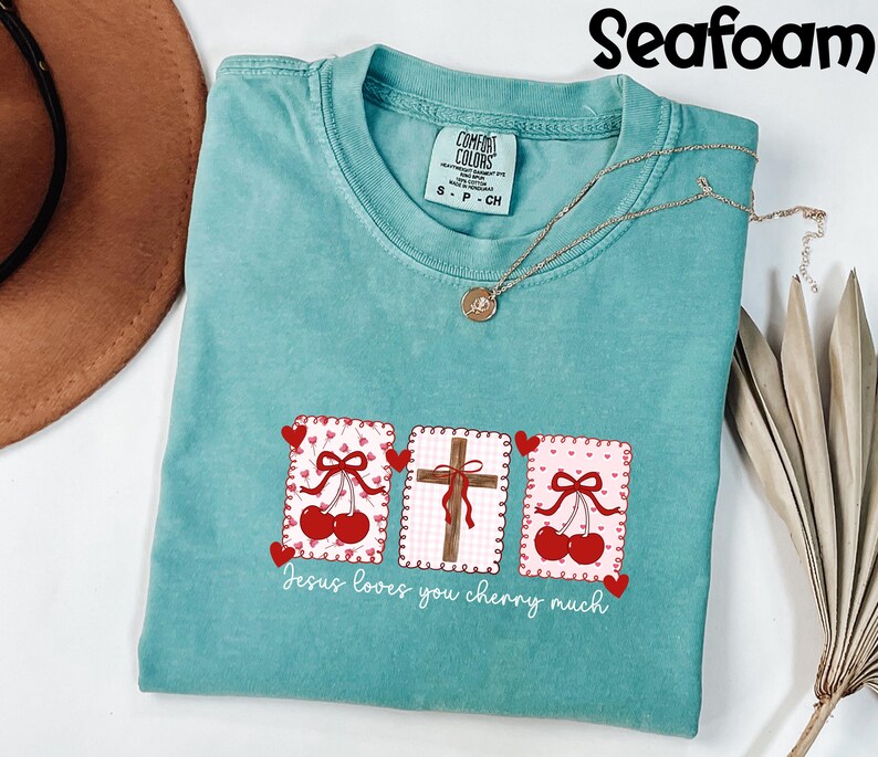 May include: Seafoam green t-shirt with a graphic design featuring cherries, a cross, and the text "Jesus loves you cherry much." The shirt has a crew neck and short sleeves. A gold necklace is draped across the shirt.