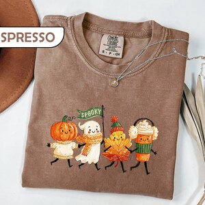 May include: An espresso-colored t-shirt featuring a graphic of a pumpkin, a ghost holding a green flag that says SPOOKY, a maple leaf, and a coffee cup, all with smiling faces. The shirt has a crew neck and a Comfort Colors brand label.