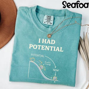 May include: Seafoam green t-shirt with the text "I HAD POTENTIAL" and a diagram illustrating potential and kinetic energy. A gold necklace with a pendant rests on the shirt. The shirt is folded on a white surface.