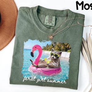 May include: Moss green t-shirt with a graphic of a raccoon wearing sunglasses and floating on a pink flamingo floatie in the ocean. The text "feral girl summer" is printed below the graphic. A gold necklace is draped on the shirt.