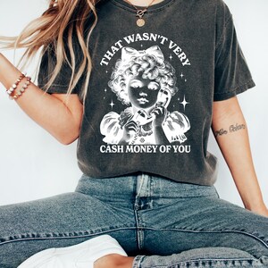 Puede incluir: Una camiseta gris oscuro con un gr&aacute;fico blanco de una ni&ntilde;a al tel&eacute;fono con el texto "That wasn't very cash money of you".