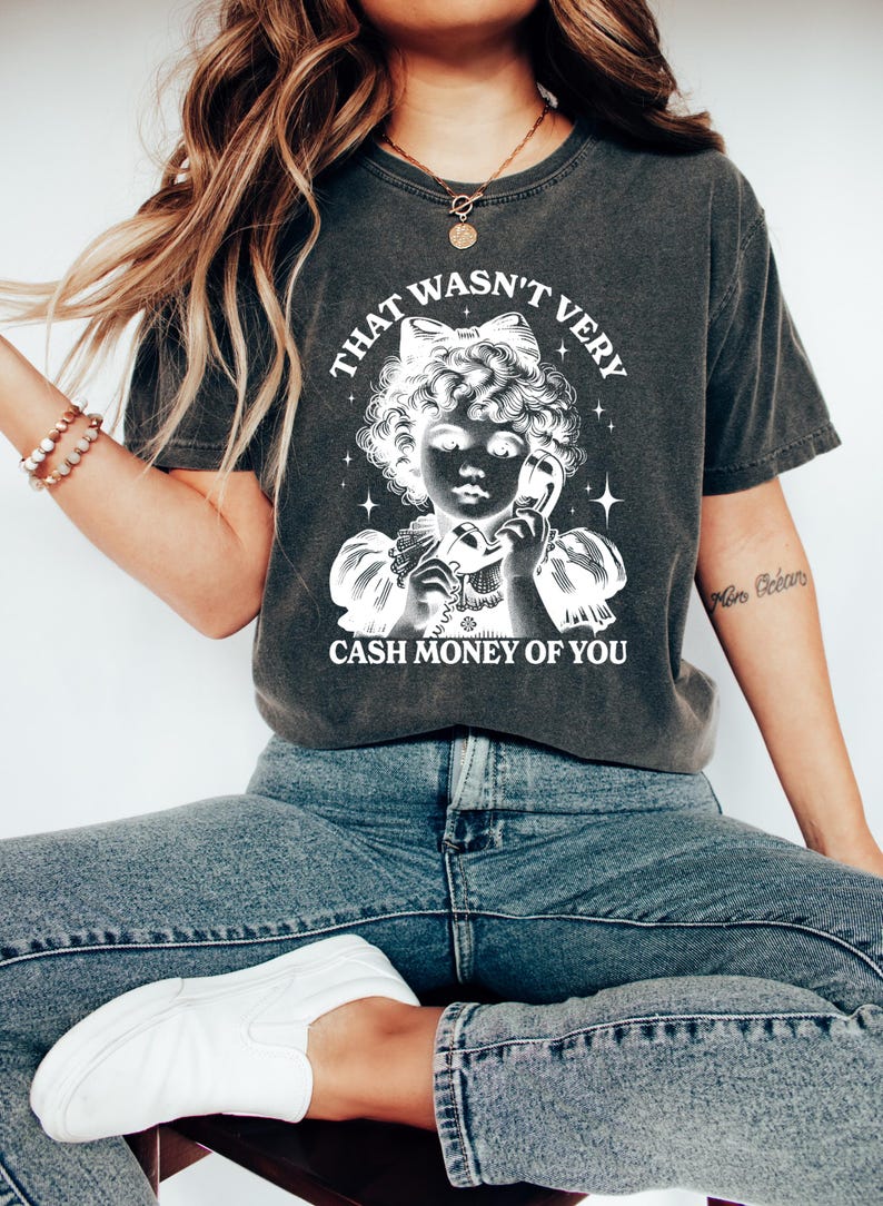 Puede incluir: Una camiseta gris oscuro con un gr&aacute;fico blanco de una ni&ntilde;a al tel&eacute;fono con el texto "That wasn't very cash money of you".