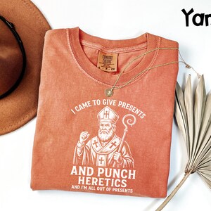 May include: A coral-coloured t-shirt with the text "I CAME TO GIVE PRESENTS AND PUNCH HERETICS AND I'M ALL OUT OF PRESENTS" and a graphic of a bishop. A brown hat and gold necklace are also in the image.