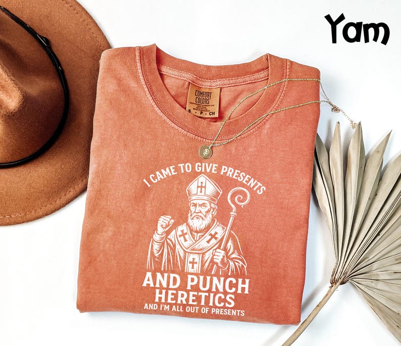 May include: A coral-coloured t-shirt with the text "I CAME TO GIVE PRESENTS AND PUNCH HERETICS AND I'M ALL OUT OF PRESENTS" and a graphic of a bishop. A brown hat and gold necklace are also in the image.