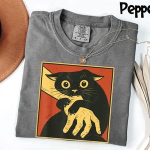 May include: Pepper-colored t-shirt featuring a graphic of a black cat biting a hand. The cat is set against a red and yellow background. The shirt has a crew neck and a Comfort Colors tag.