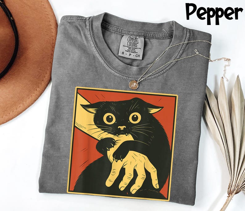 May include: Pepper-colored t-shirt featuring a graphic of a black cat biting a hand. The cat is set against a red and yellow background. The shirt has a crew neck and a Comfort Colors tag.