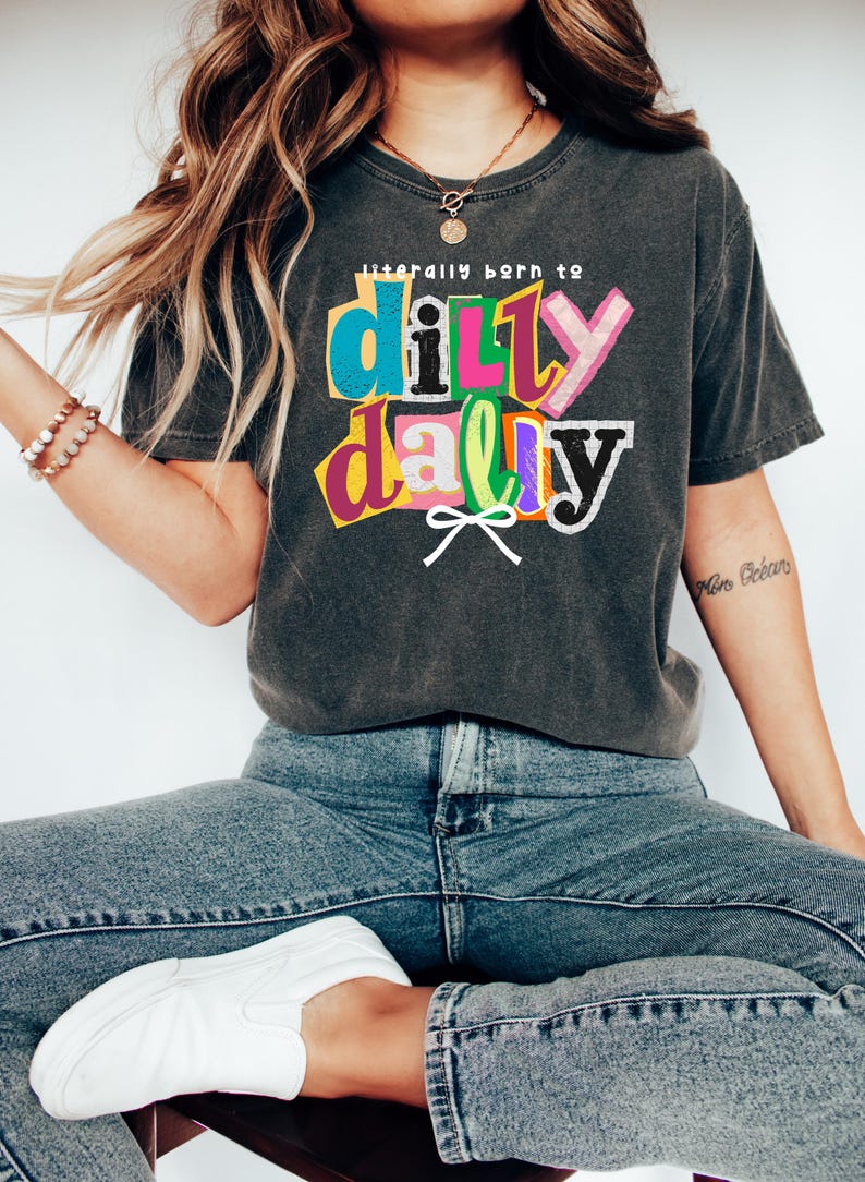 以下が含まれることがあります： カラフルな文字で「literally born to dilly dally」と書かれた、濃いグレーのTシャツ。青いジーンズと白いスリッポンシューズを合わせています。カジュアルな普段着です。