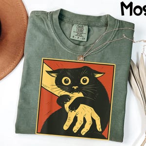 May include: A moss green t-shirt featuring a graphic of a black cat with large yellow eyes, being held by a yellow hand, set against a red background. The shirt has a Comfort Colors tag and a silver necklace.