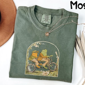 May include: A moss green t-shirt featuring a whimsical illustration of two frogs riding a tandem bicycle. The graphic is framed by an archway of flowers and foliage. The shirt has a crew neck and short sleeves.