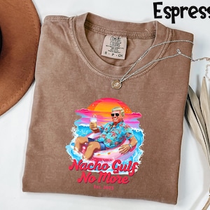 May include: Espresso-colored t-shirt with a graphic of a man in sunglasses relaxing on an inflatable ring in the ocean, holding a drink. The text "Nacho Gulf No More" is below the graphic. The shirt has a Comfort Colors tag.
