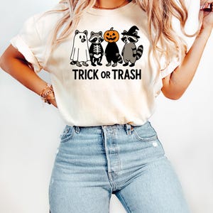 May include: Cream-colored t-shirt featuring a Halloween-themed graphic. The design includes four cartoon raccoons dressed as a ghost, skeleton, pumpkin, and witch, with the text "TRICK OR TRASH" in black.