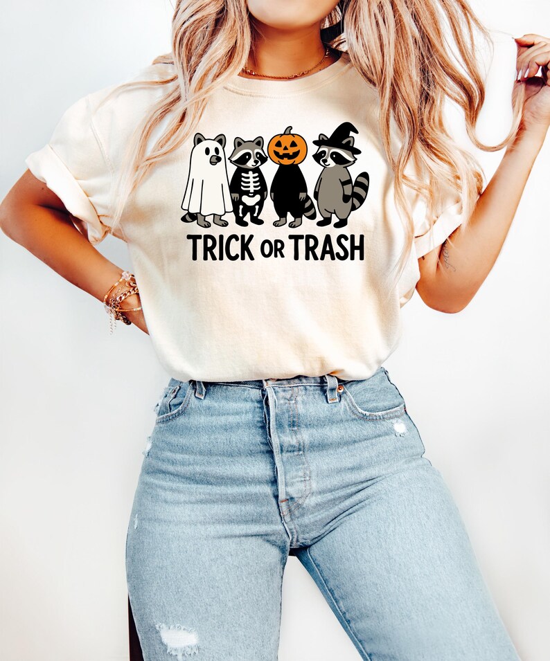 May include: Cream-colored t-shirt featuring a Halloween-themed graphic. The design includes four cartoon raccoons dressed as a ghost, skeleton, pumpkin, and witch, with the text "TRICK OR TRASH" in black.