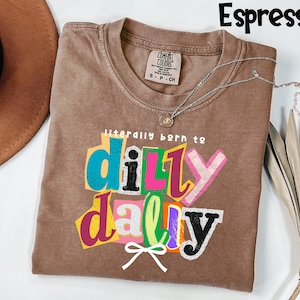 以下が含まれることがあります： エスプレッソ色のTシャツ。カラフルなレイヤーテキストで「literally born to dilly dally」というフレーズがプリントされています。Tシャツはクルーネックで、背面上部に小さなタグが付いています。ペンダント付きのシルバーネックレスがTシャツの上にかけられています。