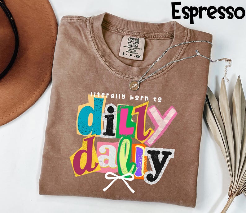 以下が含まれることがあります： エスプレッソ色のTシャツ。カラフルなレイヤーテキストで「literally born to dilly dally」というフレーズがプリントされています。Tシャツはクルーネックで、背面上部に小さなタグが付いています。ペンダント付きのシルバーネックレスがTシャツの上にかけられています。