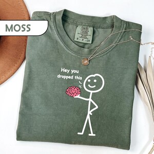 May include: A moss green short-sleeve t-shirt featuring a white stick figure graphic holding a pink brain. The text above the brain reads, "Hey you dropped this." The shirt has a crew neckline and a Comfort Colors brand label on the inside collar.