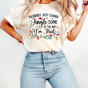 Puede incluir: Camiseta color crema con el texto "Probably Just Gonna Jingle Some of the Way I'm Tired" en negro y rojo. El dise&ntilde;o incluye un gorro de Pap&aacute; Noel, estrellas y una guirnalda de luces navide&ntilde;as de colores.