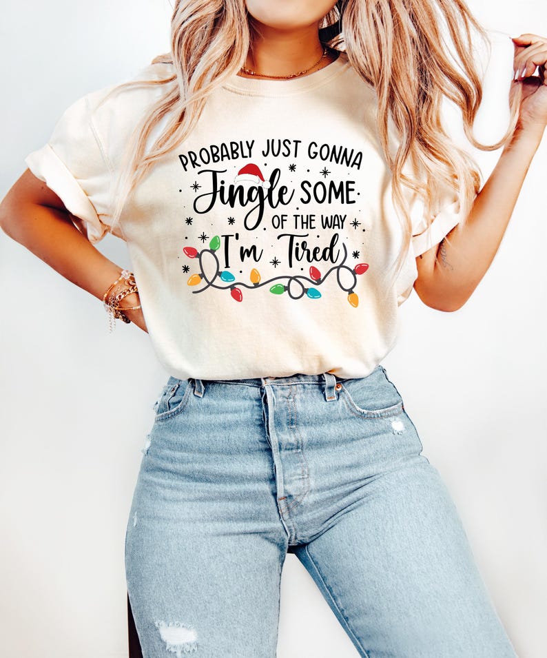Puede incluir: Camiseta color crema con el texto "Probably Just Gonna Jingle Some of the Way I'm Tired" en negro y rojo. El dise&ntilde;o incluye un gorro de Pap&aacute; Noel, estrellas y una guirnalda de luces navide&ntilde;as de colores.