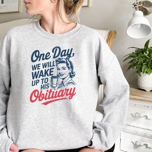 May include: Gray sweatshirt with a retro-style illustration of a woman holding a coffee cup. The text on the sweatshirt reads "One Day, We Will Wake Up To His Obituary."