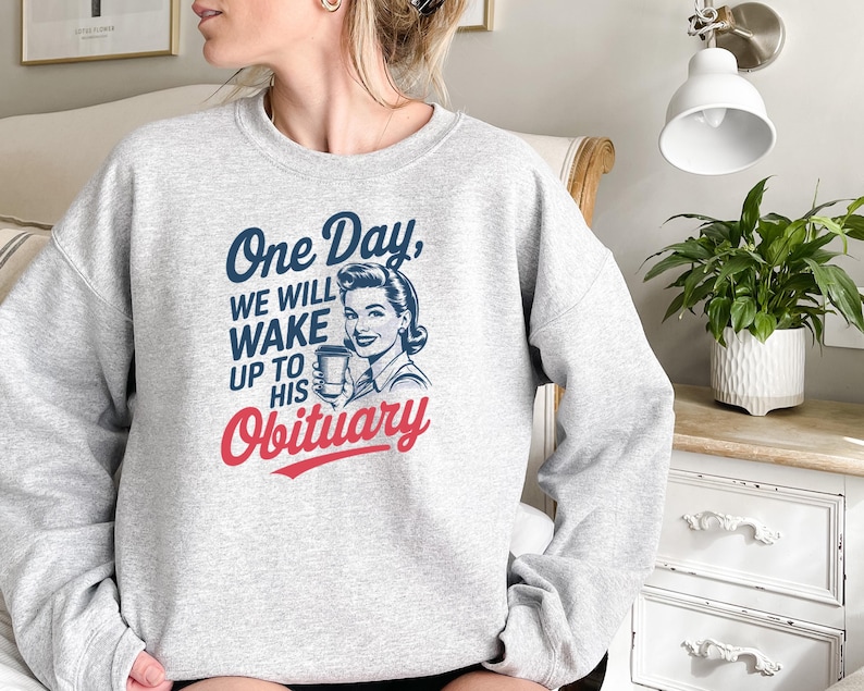 May include: Gray sweatshirt with a retro-style illustration of a woman holding a coffee cup. The text on the sweatshirt reads "One Day, We Will Wake Up To His Obituary."