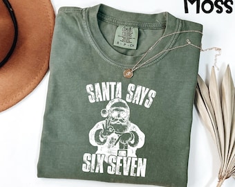 May include: A moss green t-shirt with the text "SANTA SAYS SIX SEVEN" in white. The graphic depicts Santa Claus wearing sunglasses and making a peace sign. A gold necklace with a pendant is draped on the shirt.