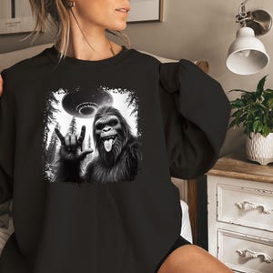 May include: Black sweatshirt with a graphic print of a Bigfoot giving the peace sign with a UFO flying overhead.
