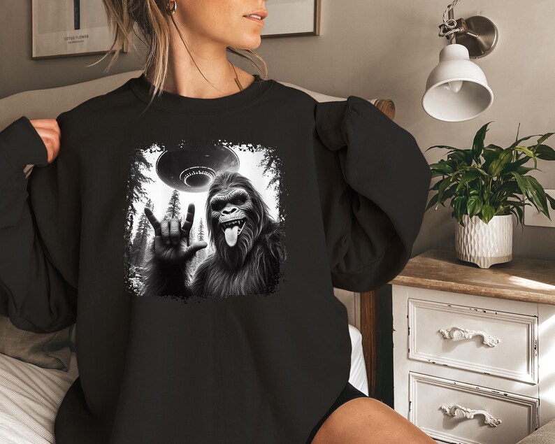 May include: Black sweatshirt with a graphic print of a Bigfoot giving the peace sign with a UFO flying overhead.