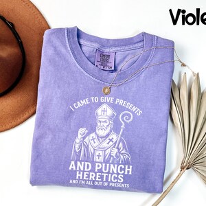 May include: A violet t-shirt with the text "I CAME TO GIVE PRESENTS AND PUNCH HERETICS AND I'M ALL OUT OF PRESENTS" and a graphic of a religious figure. A gold necklace and a brown hat are also visible.