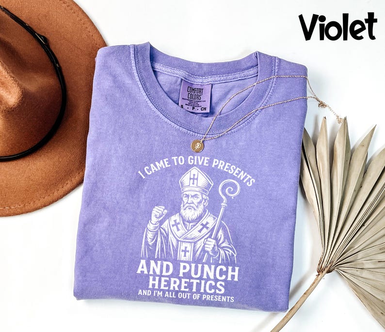 May include: A violet t-shirt with the text "I CAME TO GIVE PRESENTS AND PUNCH HERETICS AND I'M ALL OUT OF PRESENTS" and a graphic of a religious figure. A gold necklace and a brown hat are also visible.