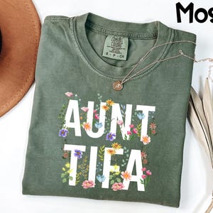 May include: A moss green t-shirt with the words "AUNT TIFIA" in white, decorated with colorful floral accents. The shirt has a crew neck and short sleeves. A gold necklace with a pendant rests on the shirt.