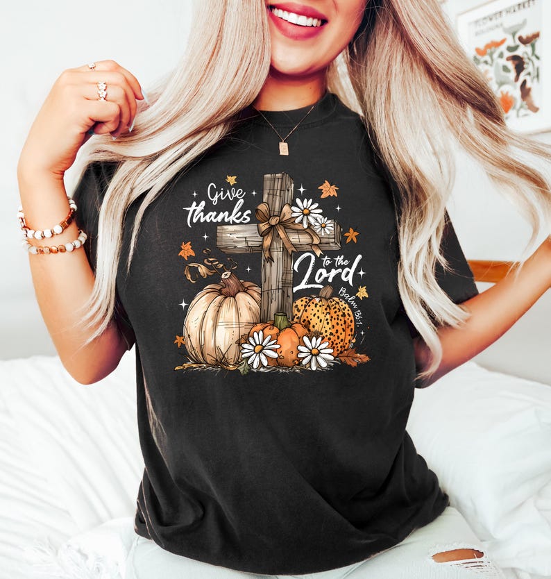 Puede incluir: Camiseta negra con un gr&aacute;fico que presenta una cruz de madera, calabazas y margaritas. El texto dice "Give thanks to the Lord". El dise&ntilde;o incluye hojas de oto&ntilde;o y un lazo, creando un tema estacional y religioso.