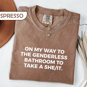 May include: An espresso-colored short-sleeve t-shirt with a crew neckline. The shirt features white text that reads: ON MY WAY TO THE GENDERLESS BATHROOM TO TAKE A SHE/IT. A silver-toned necklace with a small circular pendant rests on the fabric.