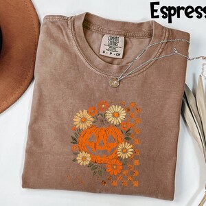 Puede incluir: Camiseta color espresso con un dise&ntilde;o de calabaza y flores. La calabaza naranja tiene una cara tallada y est&aacute; rodeada de flores naranjas y amarillas. La camiseta tiene cuello redondo y mangas cortas. Un collar plateado est&aacute; drapeado sobre la camiseta.