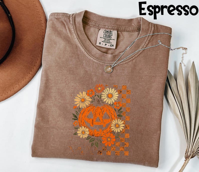 Puede incluir: Camiseta color espresso con un dise&ntilde;o de calabaza y flores. La calabaza naranja tiene una cara tallada y est&aacute; rodeada de flores naranjas y amarillas. La camiseta tiene cuello redondo y mangas cortas. Un collar plateado est&aacute; drapeado sobre la camiseta.