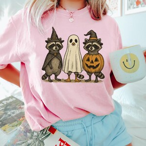 May include: Pink t-shirt featuring a Halloween-themed graphic. The graphic includes two raccoons in witch hats and capes, and a ghost. A pumpkin with a carved face is also part of the design. A white mug with a yellow smiley face is held in the image.