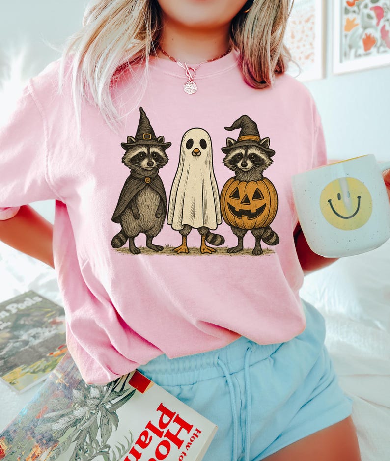 May include: Pink t-shirt featuring a Halloween-themed graphic. The graphic includes two raccoons in witch hats and capes, and a ghost. A pumpkin with a carved face is also part of the design. A white mug with a yellow smiley face is held in the image.