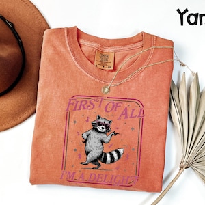 May include: A coral-coloured t-shirt featuring a raccoon in sunglasses and the text "FIRST OF ALL I'M A DELIGHT". The shirt has a crew neck and short sleeves. A gold necklace and a brown hat are also visible.