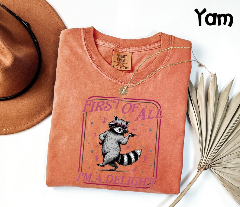May include: A coral-coloured t-shirt featuring a raccoon in sunglasses and the text "FIRST OF ALL I'M A DELIGHT". The shirt has a crew neck and short sleeves. A gold necklace and a brown hat are also visible.
