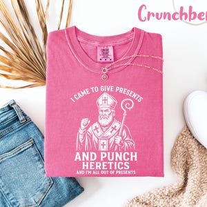 May include: A pink t-shirt with the text "I CAME TO GIVE PRESENTS AND PUNCH HERETICS AND I'M ALL OUT OF PRESENTS" and a graphic of a religious figure. The shirt is displayed with blue jeans, white shoes, and a necklace.