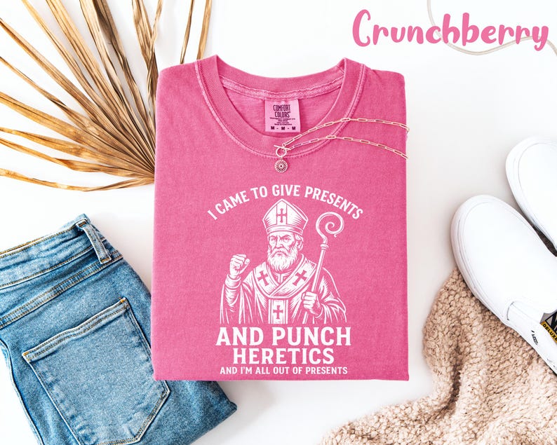May include: A pink t-shirt with the text "I CAME TO GIVE PRESENTS AND PUNCH HERETICS AND I'M ALL OUT OF PRESENTS" and a graphic of a religious figure. The shirt is displayed with blue jeans, white shoes, and a necklace.