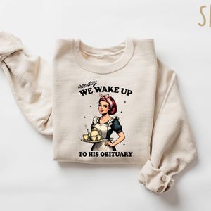 May include: A beige sweatshirt with a graphic print of a woman holding a teapot and teacups. The text on the sweatshirt reads "one day WE WAKE UP TO HIS OBITUARY".