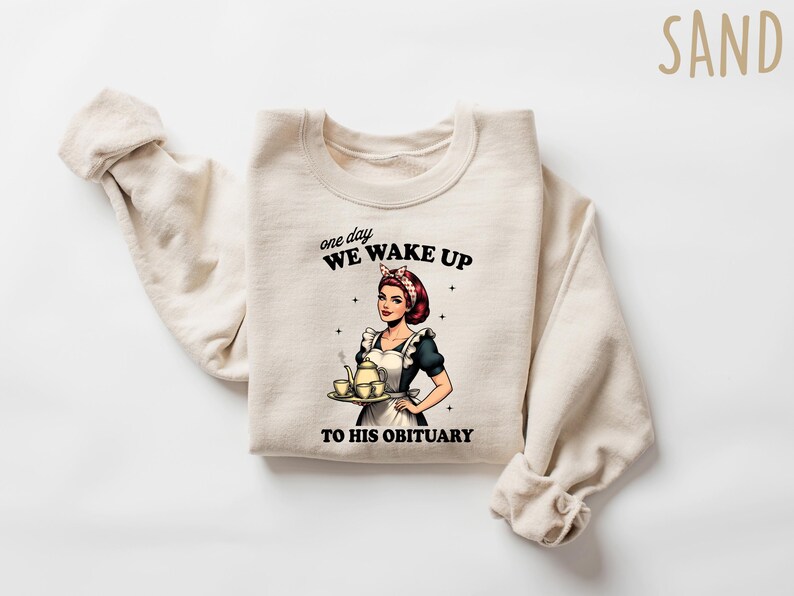 May include: A beige sweatshirt with a graphic print of a woman holding a teapot and teacups. The text on the sweatshirt reads "one day WE WAKE UP TO HIS OBITUARY".