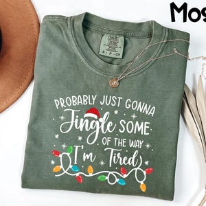 Puede incluir: Una camiseta verde musgo con el texto "Probably Just Gonna Jingle Some of the Way I'm Tired" en blanco. El dise&ntilde;o incluye un gorro de Pap&aacute; Noel y luces de cadena. Un collar dorado est&aacute; drapeado sobre la camisa.